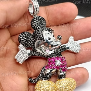 May include: A Mickey Mouse pendant with a silver bail. The pendant features black, white, pink, and gold-colored stones. The character's head and ears are black, with white gloves and gold shoes. The shorts are pink.