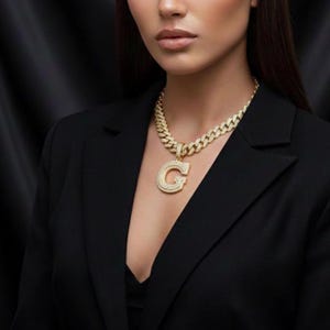 May include: A gold-colored necklace with a large pendant in the shape of the letter "G". The necklace and pendant are encrusted with small, sparkling stones. The woman is wearing a black blazer.