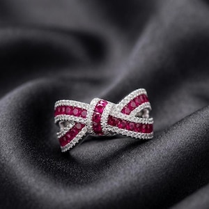 May include: A bow-shaped ring with a silver band, encrusted with small, sparkling white stones and vibrant red gemstones. The ring is set against a black satin background, highlighting its elegant design.