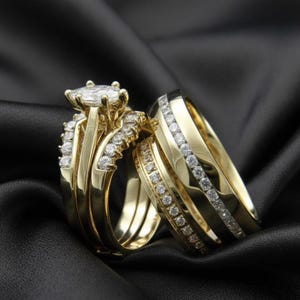 May include: A pair of gold wedding rings with diamond accents. The engagement ring features a large, oval-cut diamond center stone, and the wedding band is adorned with smaller diamonds. The rings rest on a black satin surface.