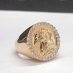 Op de afbeelding: Goudkleurige ring met een gedetailleerd muntontwerp in het midden, omringd door een halo van kleine, heldere edelstenen. De ring heeft een dikke band en een gepolijste afwerking, wat een klassieke uitstraling geeft.