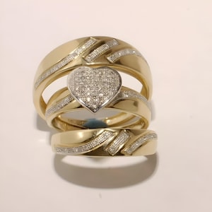 Può includere: Tre anelli d'oro, uno con un gruppo a forma di cuore di piccoli diamanti e due con una fascia di piccoli diamanti. Gli anelli sono su una superficie bianca. Gli anelli sono probabilmente per un matrimonio o un fidanzamento.