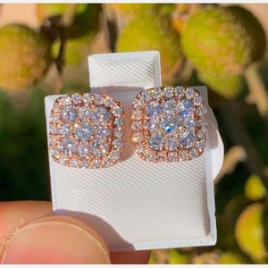 May include: A pair of square-shaped stud earrings. Each earring features a cluster of small, clear gemstones set in a rose gold-colored metal frame. The earrings are displayed on a white surface, with a blurred green background.