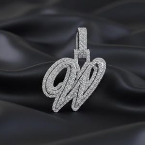 May include: A silver pendant in the shape of the letter "W", encrusted with small, sparkling stones. The pendant hangs from a matching bail, all set against a dark, silky background. The design is elegant and eye-catching.
