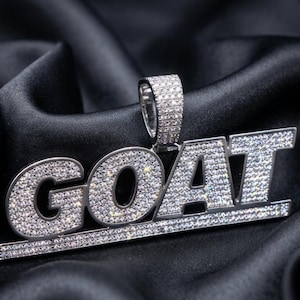 Puede incluir: Un colgante de tono plateado con la palabra GOAT en letras mayúsculas, totalmente incrustado con pequeñas piedras claras y brillantes. El colgante incluye una reasa rectangular en la parte superior, también cubierta de piedras.
