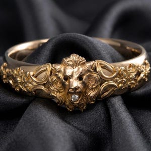 May include: Gold-toned bracelet featuring a detailed lion's head with a small diamond accent. The bracelet has an ornate, sculpted design with floral and leaf motifs. The jewelry is displayed on a dark, textured background.