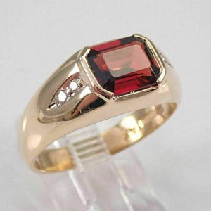 Peut inclure: Une bague en or ornée d'une grande pierre précieuse rouge foncé de forme rectangulaire. La bague a un design classique avec une bande en or poli et de petits diamants d'accent sur chaque côté de la pierre précieuse.
