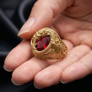 May include: A gold-toned ring featuring an intricate, textured dragon motif that wraps around a central, oval-shaped, deep red gemstone. The metalwork displays detailed scales and swirling patterns surrounding the faceted red stone setting.
