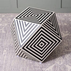 May include: Geometric cube-shaped side table with a black and white striped pattern. The cube has a series of concentric squares and diagonal stripes, creating an optical illusion. The table is set on a patterned rug.