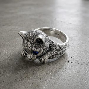 May include: A silver ring shaped like a cat with textured fur detailing and bright blue eyes. The inner band of the ring is clearly stamped with the text 925, indicating sterling silver composition. The cat design wraps around the finger.