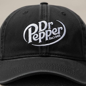 Pode incluir: Boné de beisebol preto com o logotipo da Dr Pepper bordado em branco. O logotipo apresenta as palavras "Dr Pepper" em uma fonte estilizada, com "Est. 1885" abaixo. O boné tem uma aba curva e uma alça ajustável.