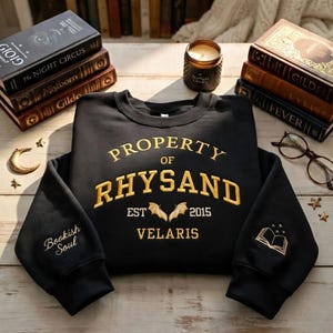 May include: Black sweatshirt with gold embroidered text reading "PROPERTY OF RHYSAND" and "VELARIS". Features a bat design and the text "EST 2015". The left sleeve says "Bookish Soul" and the right sleeve has a book design.