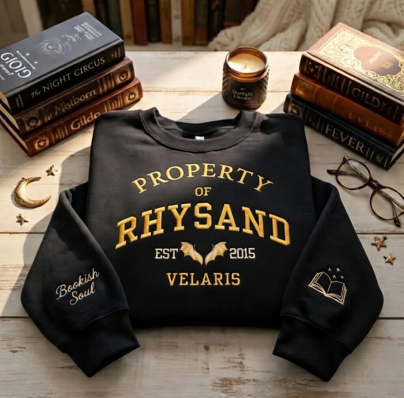 May include: Black sweatshirt with gold embroidered text reading "PROPERTY OF RHYSAND" and "VELARIS". Features a bat design and the text "EST 2015". The left sleeve says "Bookish Soul" and the right sleeve has a book design.