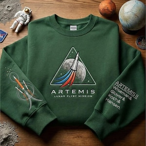 Op de afbeelding: Een donkergroene sweatshirt met het Artemis Lunar Flyby Mission logo. Het ontwerp toont een raket die achter de maan lanceert, met de tekst "ARTEMIS" en "LUNAR FLYBY MISSION". De mouw heeft de tekst "ARTEMIS II Wiseman & Glover & Koch & Hansen."