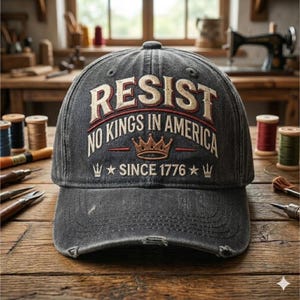 Może przedstawiać: Wypłowiała szara czapka bejsbolowa z napisem "RESIST" wyhaftowanym na biało, nad "NO KINGS IN AMERICA" i "SINCE 1776". Czapka posiada koronę i gwiazdki, z postarzanym wyglądem.