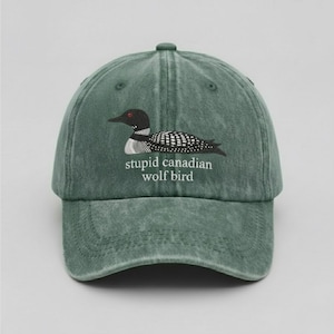 May include: A green baseball cap with a distressed finish. The cap features an embroidered design of a loon with the text "stupid canadian wolf bird" in white. The loon has a black head, red eye, and a white and black patterned body.