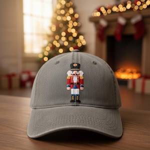 Könnte beinhalten: Graue Baseballkappe mit einem gestickten Nussknacker-Design. Der Nussknacker ist in traditionellen Farben gehalten: Rot, Blau, Weiß und Gold. Die Kappe ist vor einem festlichen Hintergrund platziert, was auf ein saisonales Thema hindeutet.