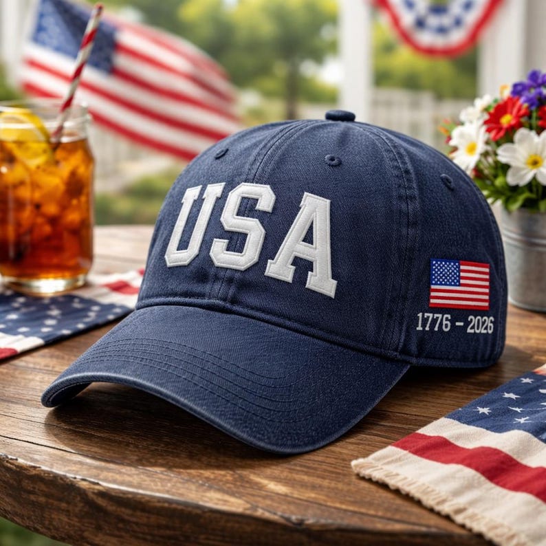 May include: A navy blue baseball cap with the word "USA" in white block letters. The cap features a small American flag and the dates "1776-2026" on the side. The cap is sitting on a wooden table, with a glass of iced tea and an American flag in the background.