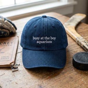 May include: A navy blue baseball cap with the text "busy at the boy aquarium" embroidered in white. The cap is on a wooden surface with hockey-related items, including a hockey stick and puck.