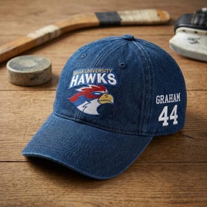 Op de afbeelding: Een marineblauwe denim baseballcap met de tekst "Briar University Hawks" en een havikenlogo. De naam "Graham" en het nummer "44" staan op de zijkant. Hockeyuitrusting op de achtergrond.