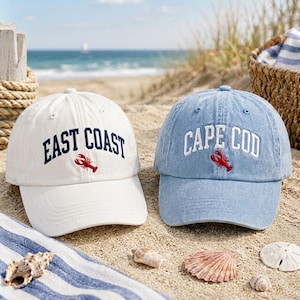 Könnte beinhalten: Zwei Baseballkappen, eine weiße mit dunkelblauem Schriftzug EAST COAST und eine hellblaue mit weißem Schriftzug CAPE COD, beide mit einer gestickten roten Hummergrafik, die unter dem Text auf den Vorderteilen zentriert ist.