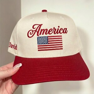 Könnte beinhalten: Cremefarbene und rote Baseballkappe mit dem Wort "America" in roter Schrift gestickt, zusammen mit einer gestickten amerikanischen Flagge. Der Schirm ist rot, und der Name "David" ist auf der Seite gestickt.