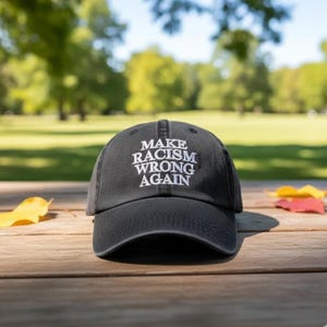 May include: A black baseball cap with white embroidered text reading "MAKE RACISM WRONG AGAIN". The cap is on a wooden surface, with a blurred background of trees and greenery.