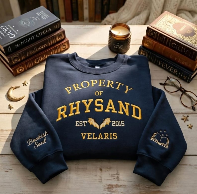 May include: Navy blue sweatshirt with gold embroidered text reading "PROPERTY OF RHYSAND EST 2015 VELARIS." The left sleeve says "Bookish Soul" and the right sleeve has a book graphic. Books and a candle are in the background.