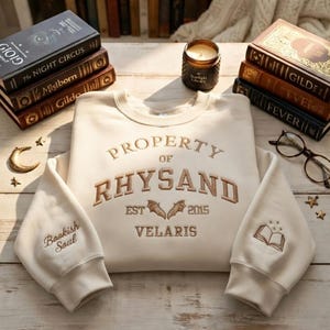May include: Cream-colored sweatshirt with embroidered text: "PROPERTY OF RHYSAND EST 2015 VELARIS." Features bats and a book graphic. Sleeves have "Bookish Soul" and a book graphic. Books, a candle, and glasses are in the background.