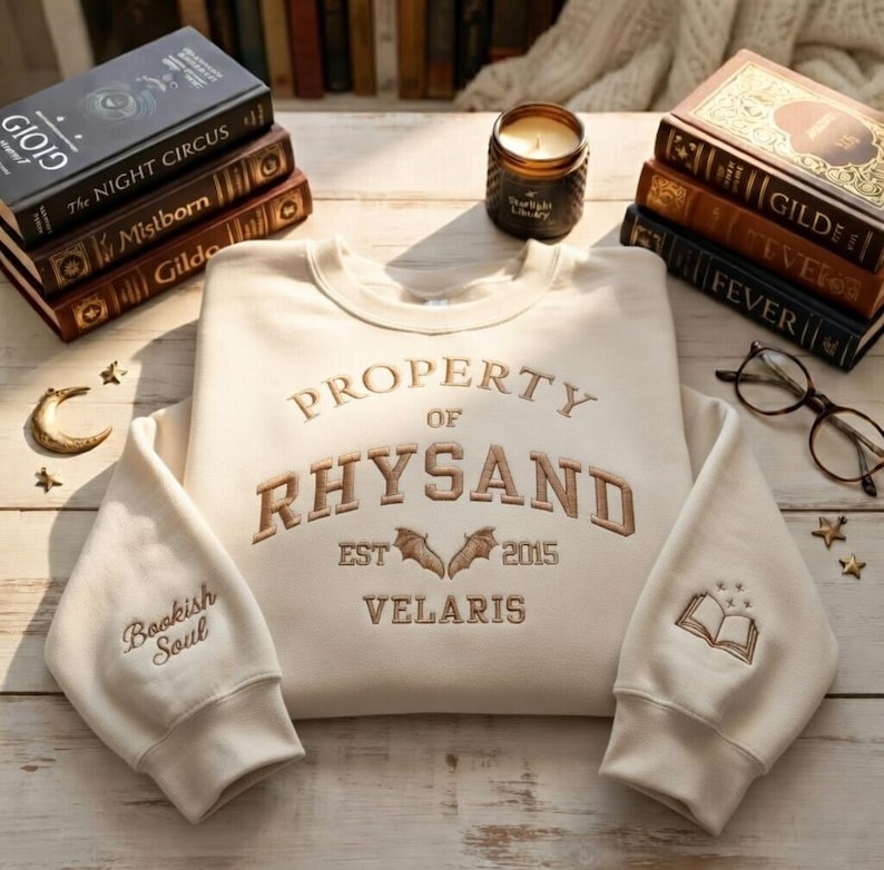 May include: Cream-colored sweatshirt with embroidered text: "PROPERTY OF RHYSAND EST 2015 VELARIS." Features bats and a book graphic. Sleeves have "Bookish Soul" and a book graphic. Books, a candle, and glasses are in the background.