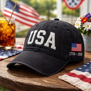 May include: Black baseball cap with the word "USA" in white, outlined letters. The cap features a small American flag and the years "1756-2026" on the side. The cap is on a wooden table with an American flag in the background.