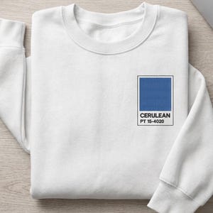May include: White crew neck sweatshirt with a small square graphic of cerulean blue color and the text "CERULEAN PT 15-4020". The sweatshirt has long sleeves and a relaxed fit.
