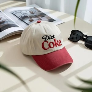 May include: A cream-coloured baseball cap with a red brim and button. The words "Diet Coke" are embroidered in black script with red lettering. Black sunglasses are to the right of the hat.
