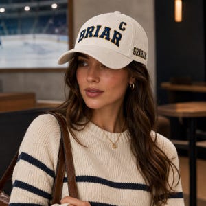 May include: A cream-colored baseball cap with the word "BRIAR" arched across the front in navy blue letters. The letter "C" is above the word "GRAHAM" on the right side. The cap has a curved brim.