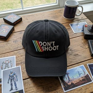 May include: Black baseball cap with the text "DON'T SHOOT" in white, with a colorful striped design. The cap is on a wooden surface with other items, including a mug and game cards.