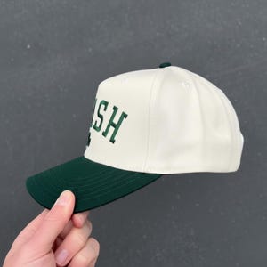 Peut inclure: Casquette de baseball blanche et verte avec le mot "IRISH" brod&eacute; en lettres vertes. La casquette a une visi&egrave;re verte et un bouton sur le dessus. Le chapeau est de style classique.