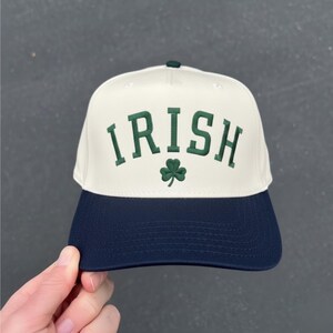 Peut inclure: Casquette de baseball blanche et bleu marine avec le mot "IRISH" brod&eacute; en vert, ainsi qu'un tr&egrave;fle vert. La casquette a une visi&egrave;re incurv&eacute;e et un bouton sur le dessus.