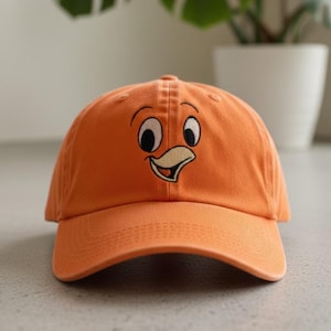 Puede incluir: Gorra de béisbol naranja con un rostro de pájaro de dibujos animados bordado. El diseño incluye ojos negros, un pico amarillo y una boca sonriente. La gorra tiene una visera curva y un cierre ajustable. Un accesorio divertido e informal.