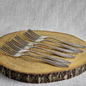 Puede incluir: Seis tenedores plateados con diseños florales ornamentados están colocados sobre una tabla redonda de madera. Los tenedores están sobre una superficie de madera clara y texturizada con borde de corteza natural. El fondo es de tela de lino gris claro.