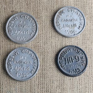 Peut inclure: Quatre plaques métalliques circulaires avec du texte cyrillique en relief. Trois plaques sont argentées et datées de 1966, avec le texte "ПЕРВЫЙ СИЛУМИН ИЗ КАОЛИНА АМЕТИ 1966". Une plaque est gris foncé, datée de 1974, avec "ПЕРВЫЙ ЧУГУН ДП-9 31XII1974 КМЗ".
