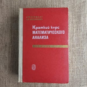 Może przedstawiać: Czerwona książka z białym tekstem, zatytułowana "Kratkii kurs Matematicheskogo Analiza" w języku rosyjskim. Książka ma złoty poziomy pasek i nazwiska autorów na górze. Książka leży na teksturowanej, beżowej powierzchni.