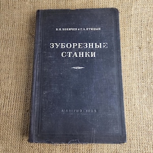 Könnte beinhalten: Ein dunkelblaues, vintage Buch mit dem Titel "ЗУБОРЕЗНЫЕ СТАНКИ" in weißer kyrillischer Schrift. Das Cover zeigt auch die Namen "В.Н. КОКИЧЕВ И Г.А.ПТИЦЫН" und das Jahr "1954". Das Buch liegt vor einem strukturierten, beigefarbenen Hintergrund.