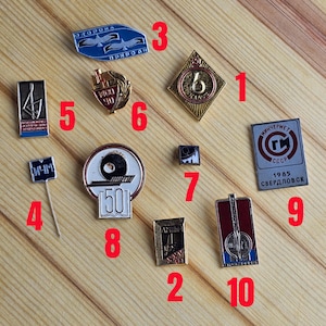 May include: A collection of vintage metal pins and badges, featuring various designs and inscriptions. The pins include a diamond-shaped badge with the number 6, a blue badge with birds, and a pin with the text "1985 Sverdlovsk".