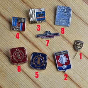Pode incluir: Uma coleção de alfinetes de metal vintage com vários designs. Os alfinetes apresentam imagens de um farol, um edifício, uma bandeira e outros símbolos. Alguns alfinetes têm texto, incluindo "200" e outras palavras.