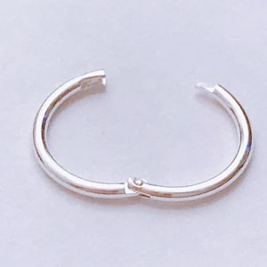 May include: A silver hinged hoop earring, open and ready to be worn. The earring is a simple, circular design with a clasp mechanism. The metal has a polished, reflective surface, and the earring is set against a light background.