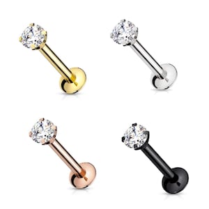 May include: Four labret studs featuring a round, clear, faceted gemstone held by four prongs. The posts and flat back discs are available in gold, silver, rose gold, and black metal finishes, each displayed at an angle to show the structure.