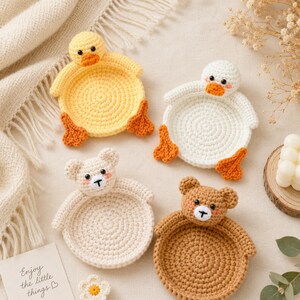May include: Four round crochet coasters featuring animal designs. Two are yellow and white ducks with orange beaks and feet, and two are beige and brown bears with pink cheeks. Each coaster has a circular base and a small animal head at the top.