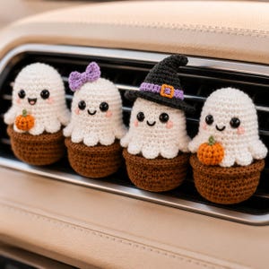May include: Four small crocheted ghost figures in brown pots, each with black eyes and pink cheeks. One ghost has a small orange pumpkin, one has a purple bow, one wears a black witch hat with a purple band, and one holds an orange pumpkin.