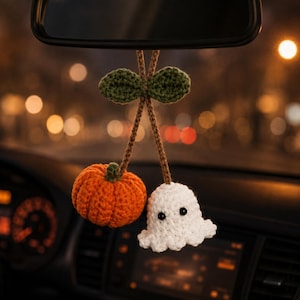 May include: A crochet car accessory featuring an orange pumpkin and a white ghost hanging from brown yarn stems with two green leaves at the top. The ghost has two small black bead eyes and a scalloped bottom edge, while the pumpkin is ribbed.