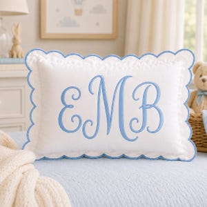 May include: A rectangular white pillow featuring a scalloped edge trimmed in light blue thread. The center of the pillow displays the embroidered monogram letters E, M, and B in a large, cursive, light blue font with decorative flourishes.